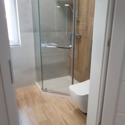 Salbox - Nowoczesna łazienka z kabiną prysznicową typu walk-in, drewnopodobną podłogą i podwieszaną toaletą, widok zza częściowo otwartych białych drzwi.