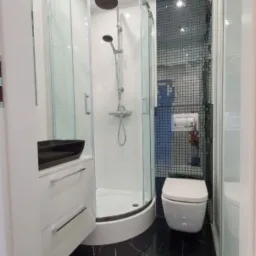 Nowoczesna łazienka z czarną marmurową podłogą, kabiną prysznicową narożną z przezroczystego szkła, białą umywalką nablatową i toaletą w zabudowie na tle ściany wyłożonej mozaiką.