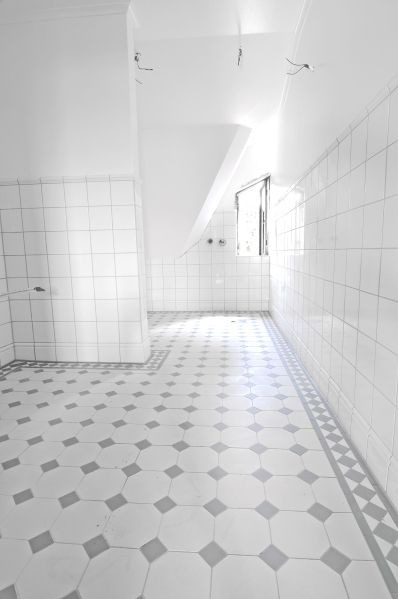 Wnętrze łazienki po remoncie z białymi płytkami na ścianach i podłodze w geometryczne wzory, widoczne przewody elektryczne wystające z sufitu.