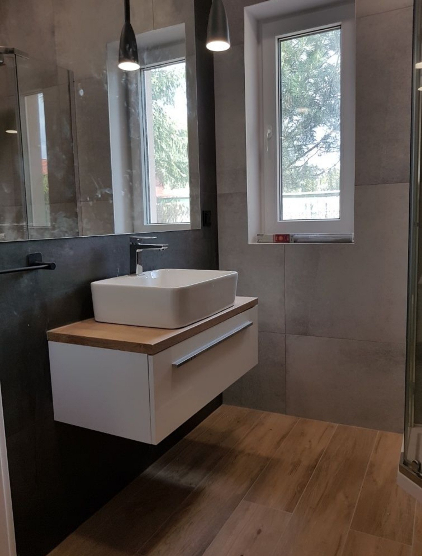 Nowoczesna łazienka z wiszącą szafką pod umywalką nablatową, drewnianym blatem i szarą płytką imitującą kamień na ścianach, oświetlona wiszącymi lampami i naturalnym światłem z okna.