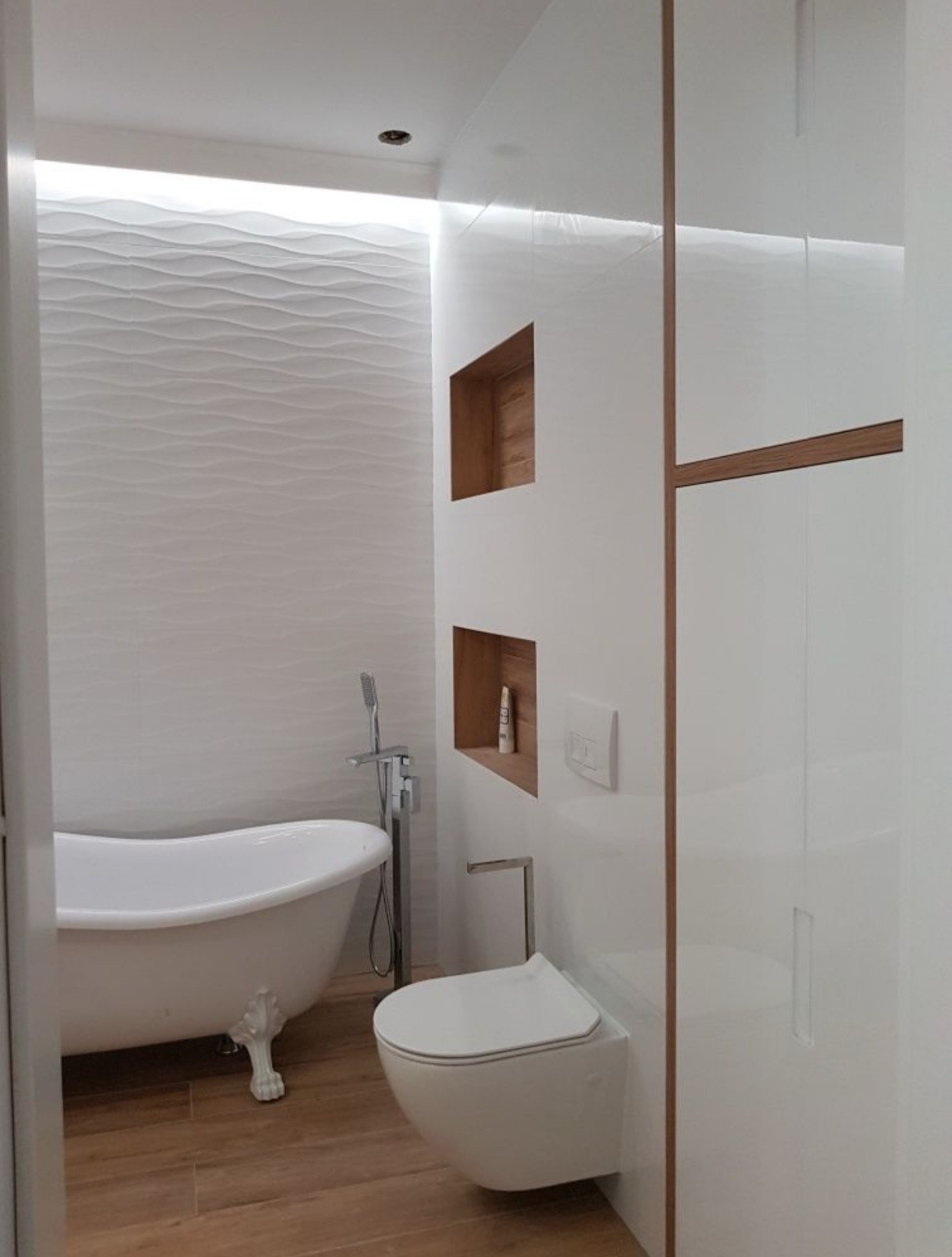 Nowoczesna łazienka z białą wanną na lwich nóżkach, toaletą podwieszaną i ścianą z trójwymiarowych białych płytek, oświetlona listwami LED.