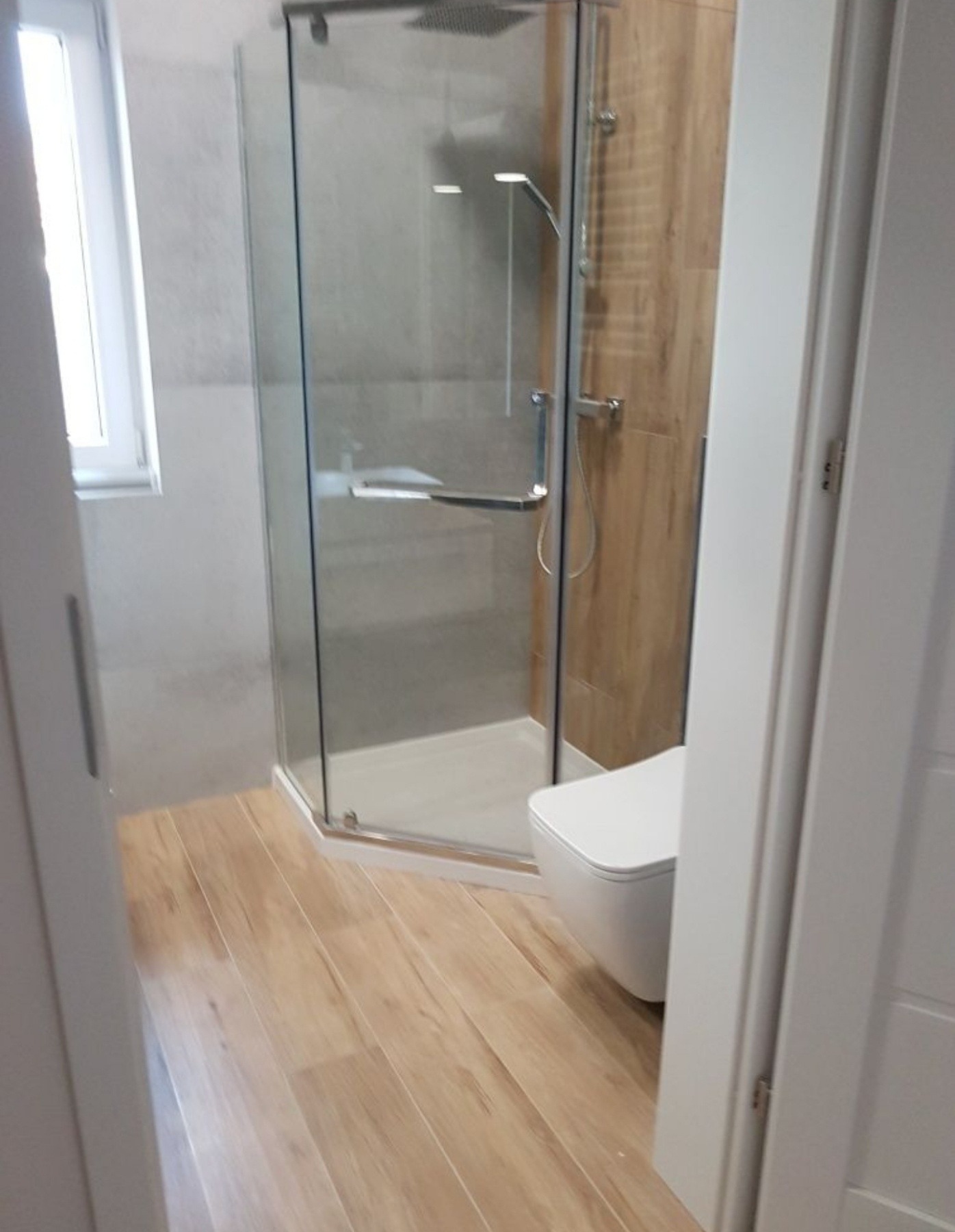 Nowoczesna łazienka z kabiną prysznicową typu walk-in, drewnopodobną podłogą i podwieszaną toaletą, widok zza częściowo otwartych białych drzwi.