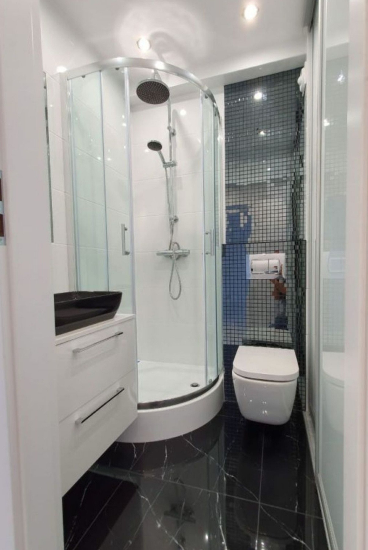 Nowoczesna łazienka z czarną marmurową podłogą, kabiną prysznicową narożną z przezroczystego szkła, białą umywalką nablatową i toaletą w zabudowie na tle ściany wyłożonej mozaiką.