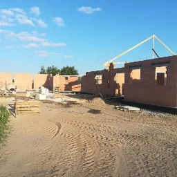 Wznoszenie ścian parterowego domu z czerwonej cegły, widoczne otwory okienne i początek konstrukcji dachu, na placu budowy leży beton i stoi betoniarka.