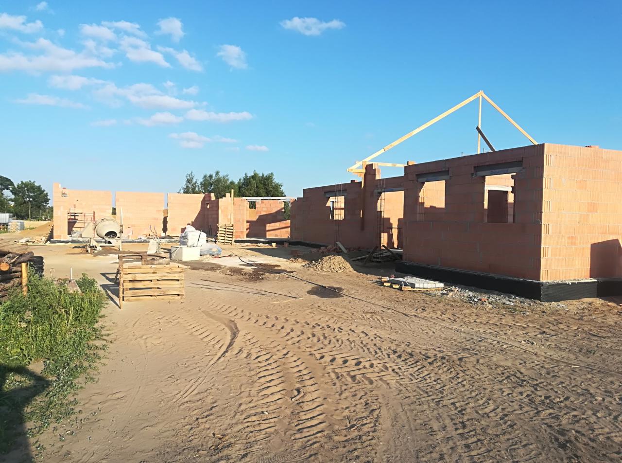 Wznoszenie ścian parterowego domu z czerwonej cegły, widoczne otwory okienne i początek konstrukcji dachu, na placu budowy leży beton i stoi betoniarka.