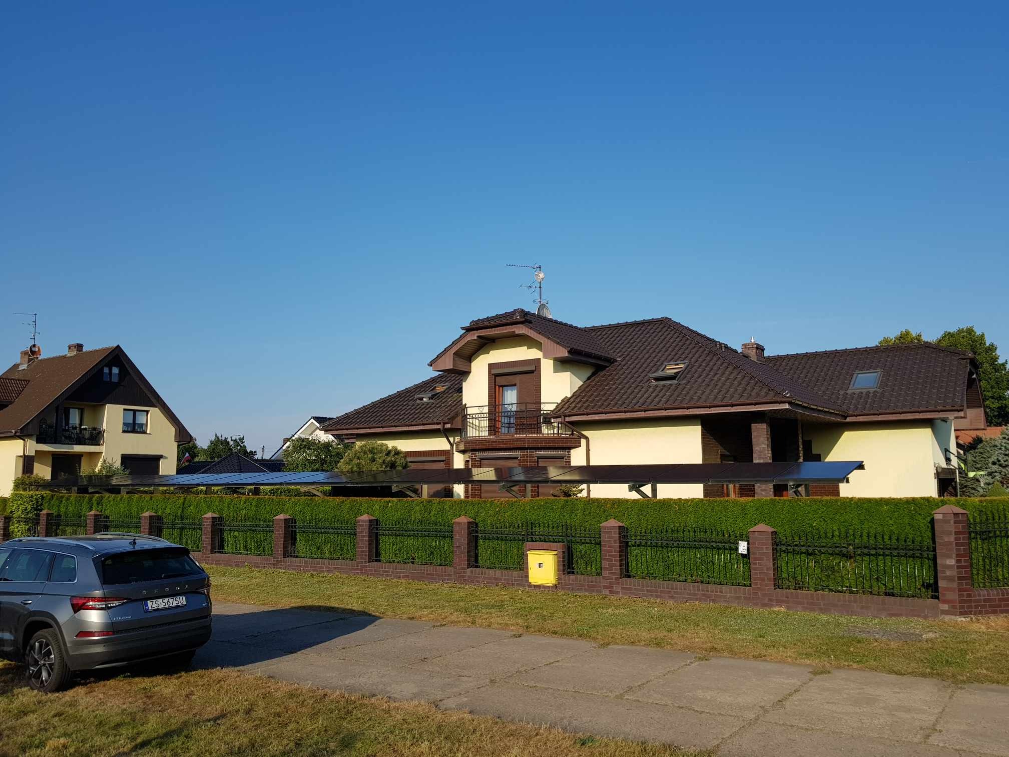 Dom jednorodzinny z panelami słonecznymi widocznymi za żywopłotem i ogrodzeniem. Szary samochód osobowy zaparkowany z boku. Błękitne niebo w tle.