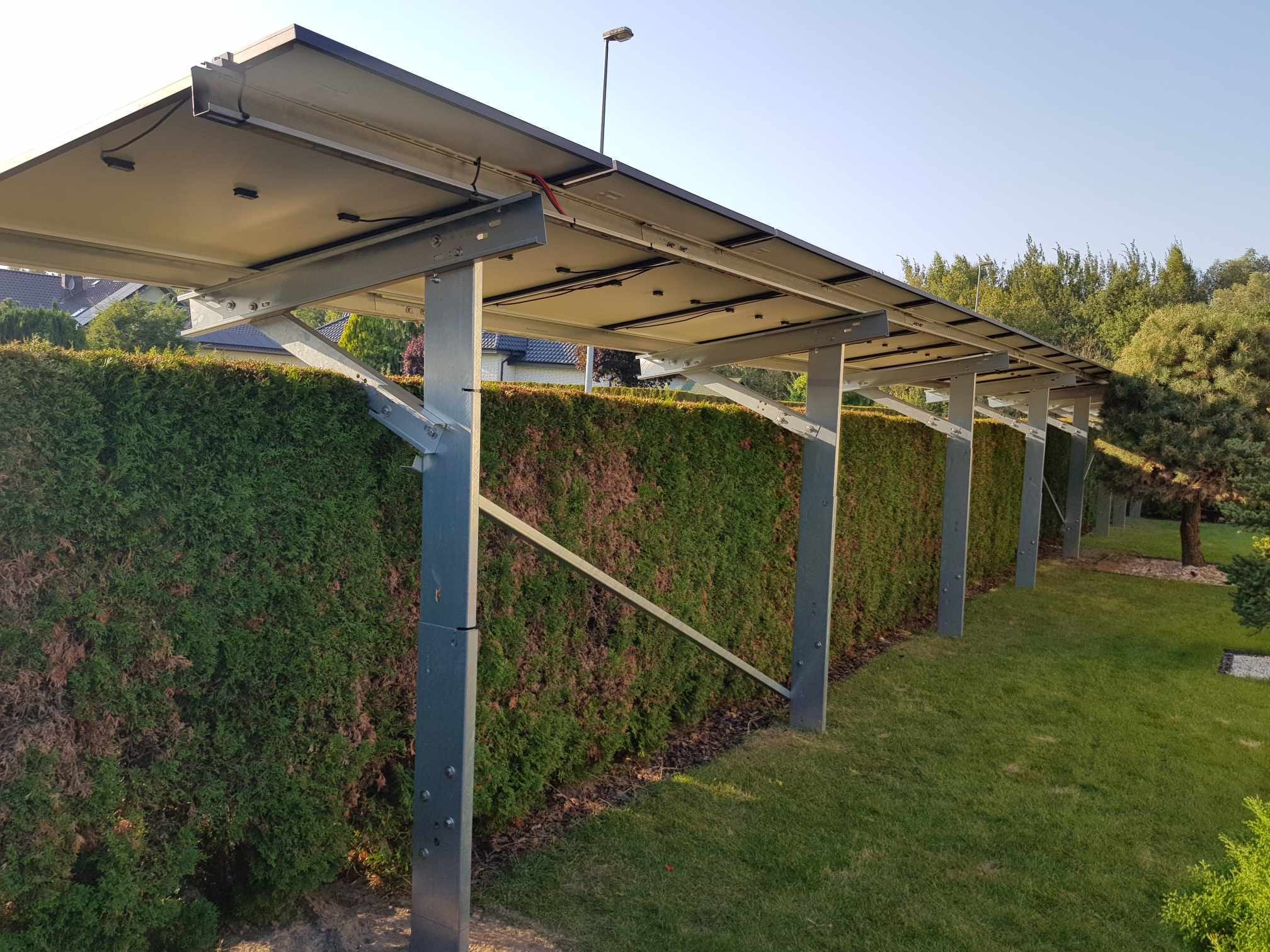 Zadaszenie fotowoltaiczne nad żywopłotem, wsparte na metalowych słupach. Widoczne panele od spodu, instalacja na tle zieleni i błękitnego nieba.