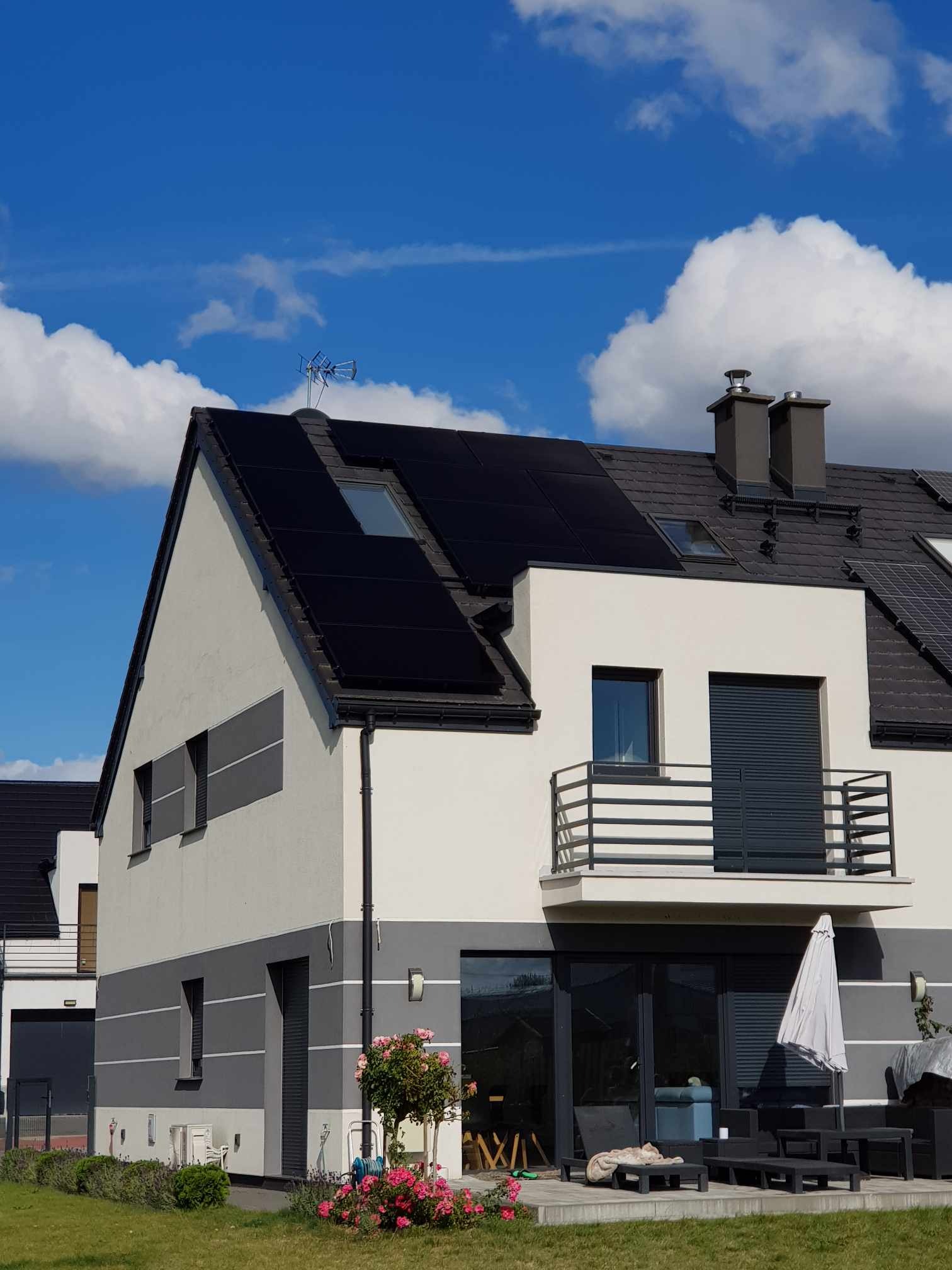 Dwukondygnacyjny dom z panelami słonecznymi na dachu, widoczne z poziomu ogrodu z kwiatami i meblami tarasowymi. Antena na dachu. Błękitne niebo z chmurami w tle.