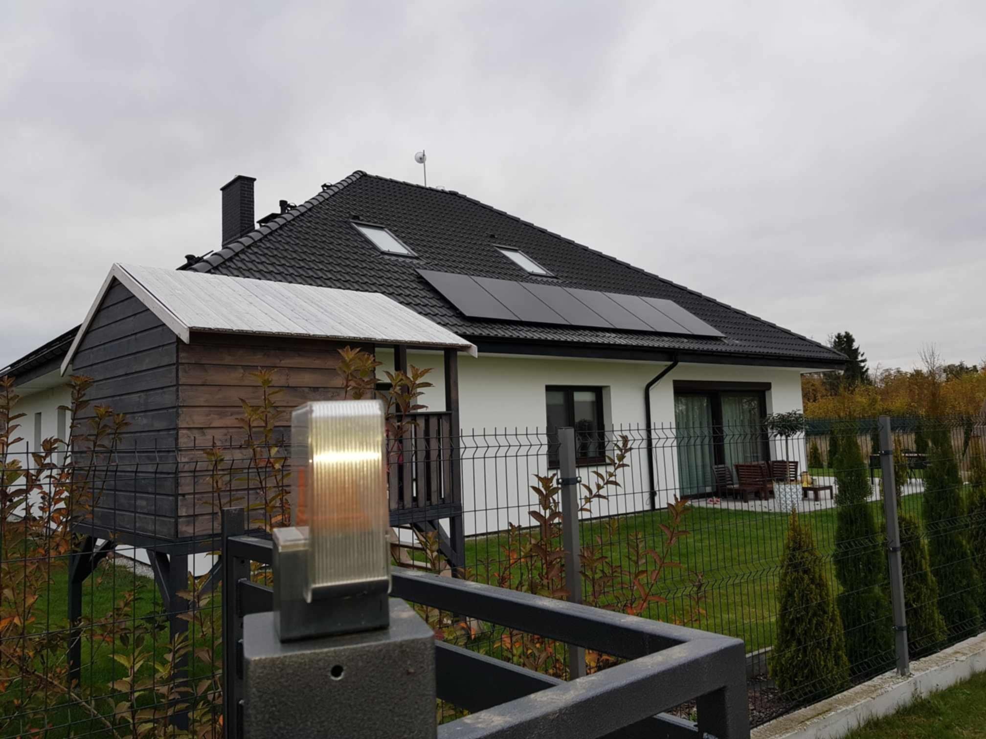 Dom jednorodzinny z czarnym dachem i panelami słonecznymi, widoczny zza ogrodzenia. Na pierwszym planie lampa ogrodowa i drewniany domek na podwyższeniu.