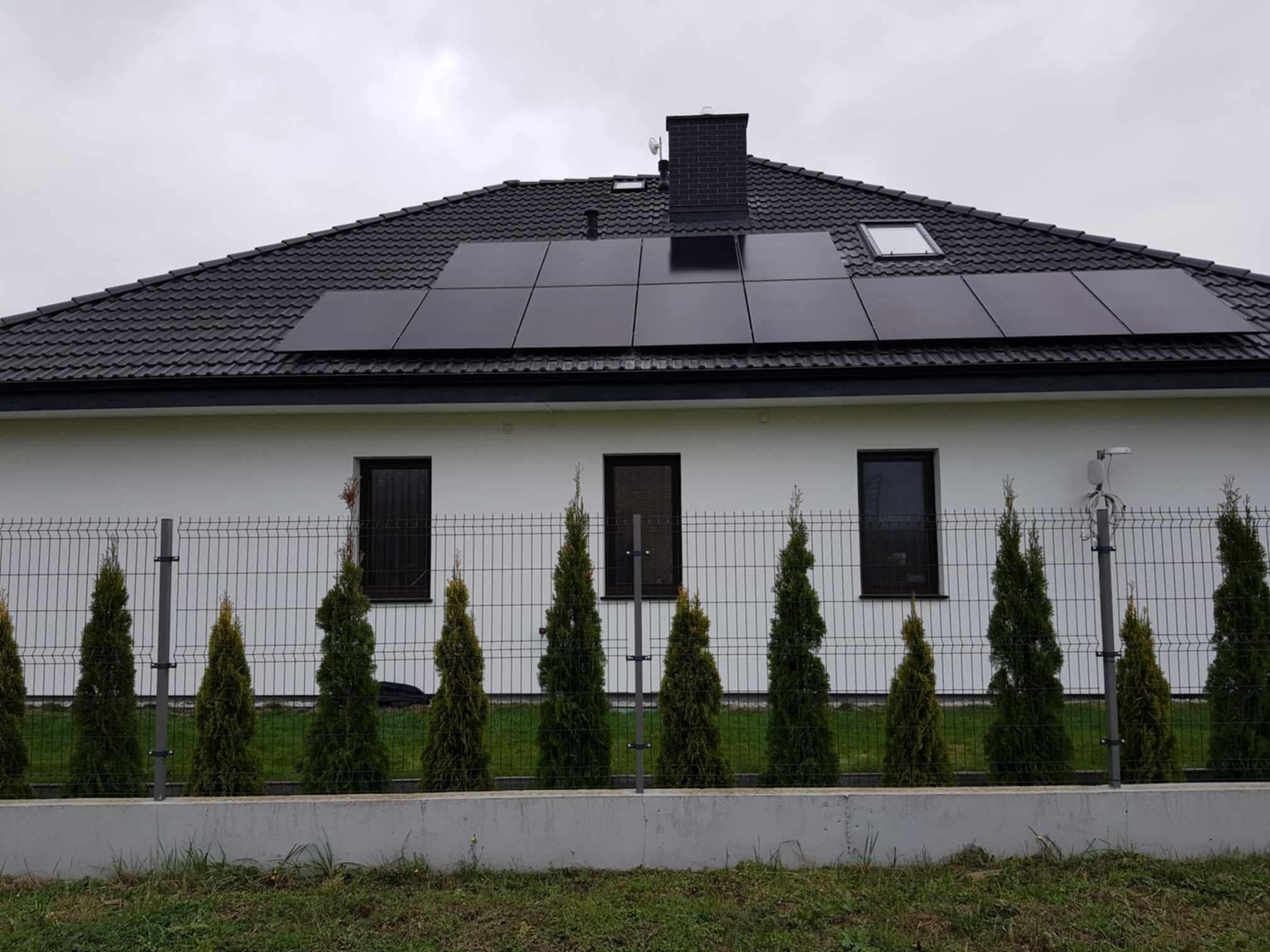 Czarny dach z panelami słonecznymi na białym domu jednorodzinnym, widok zza ogrodzenia z tujami. Pochmurne niebo.