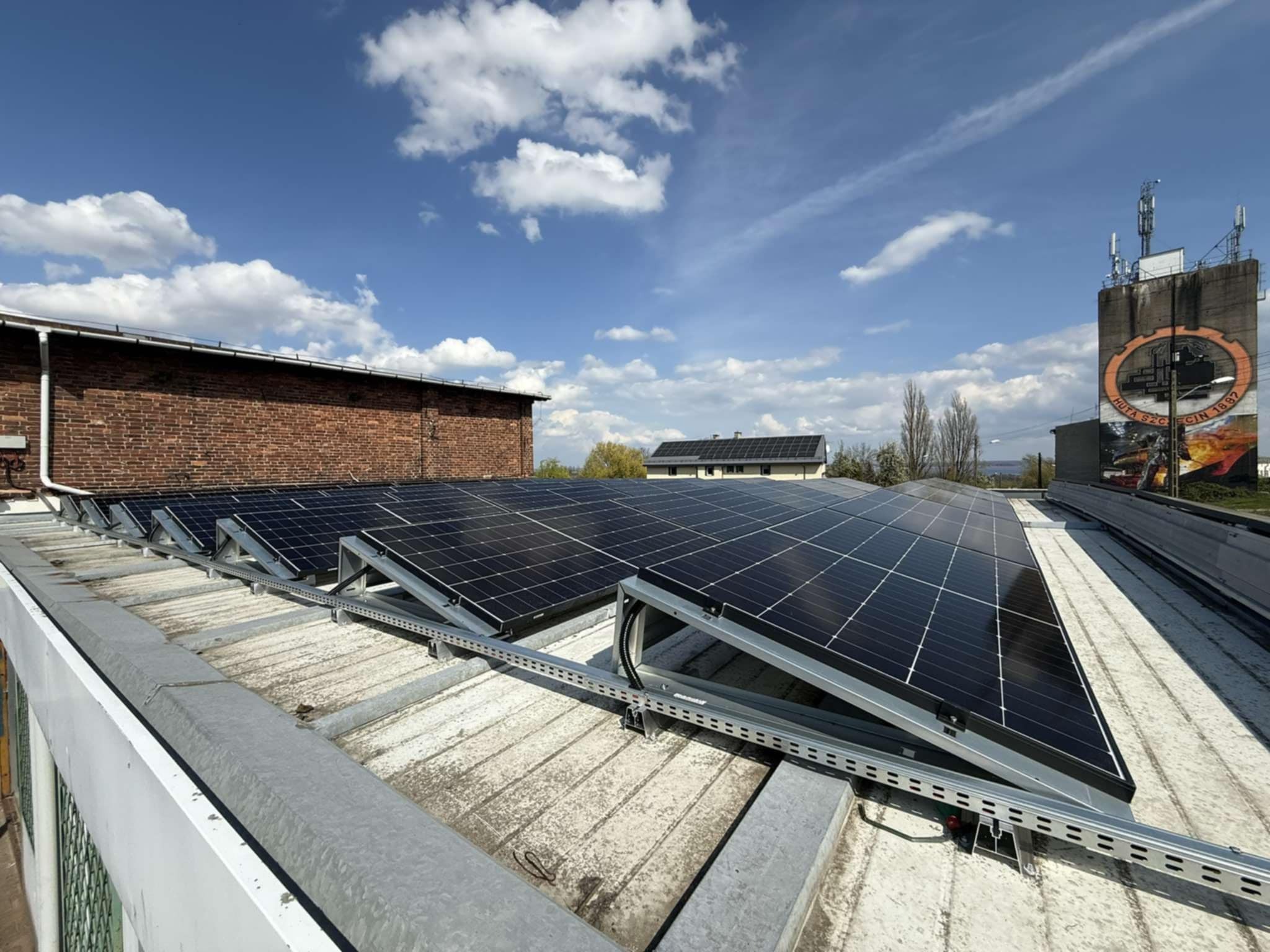 Panele słoneczne na dachu budynku z widoczną konstrukcją montażową, w tle budynek z muralem i błękitne niebo z chmurami.