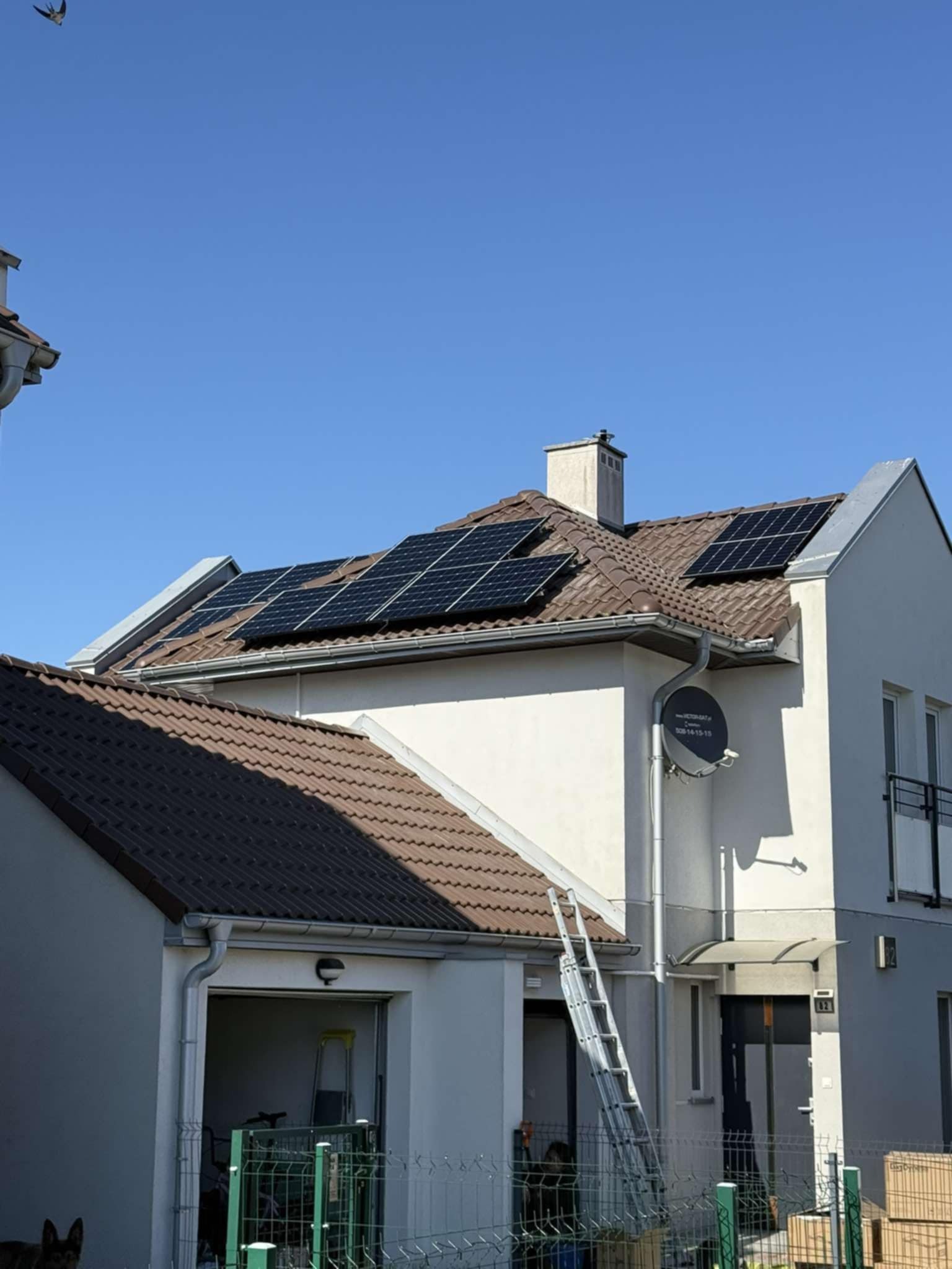 Biały dom z brązowym dachem i panelami słonecznymi. Aluminiowa drabina oparta o rynnę. Antena satelitarna na ścianie. Błękitne niebo w tle.