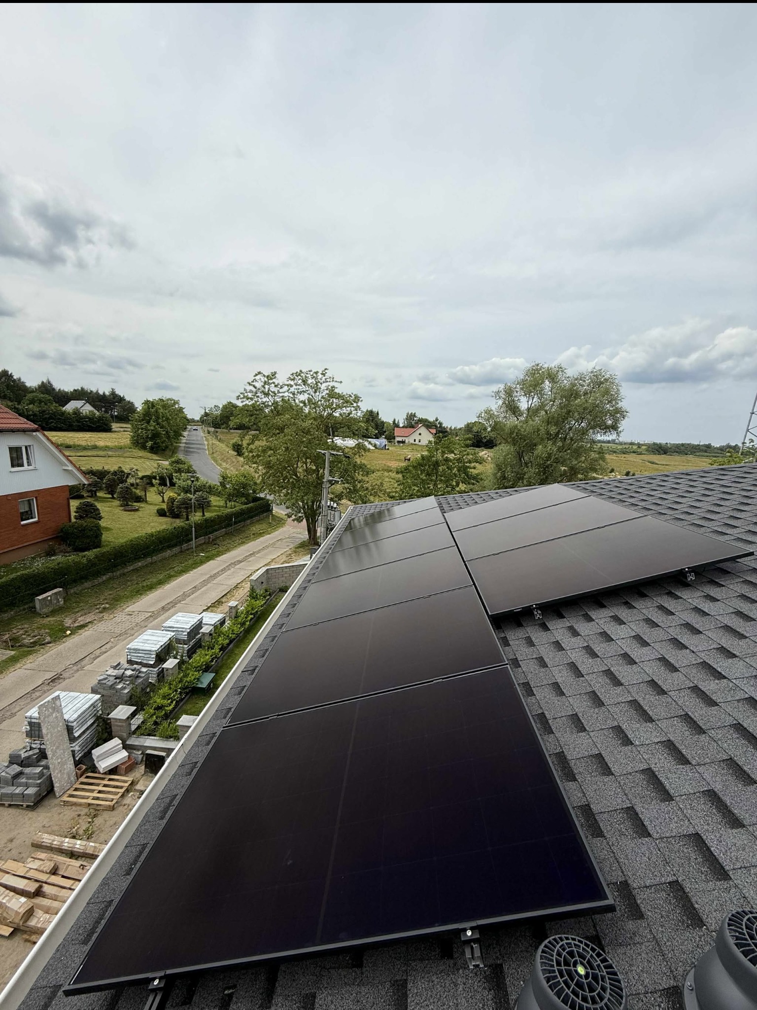 Cztery czarne panele fotowoltaiczne na dachu pokrytym gontem bitumicznym, widok z góry na okolicę z domami i drogą w pochmurny dzień. Montaż paneli.