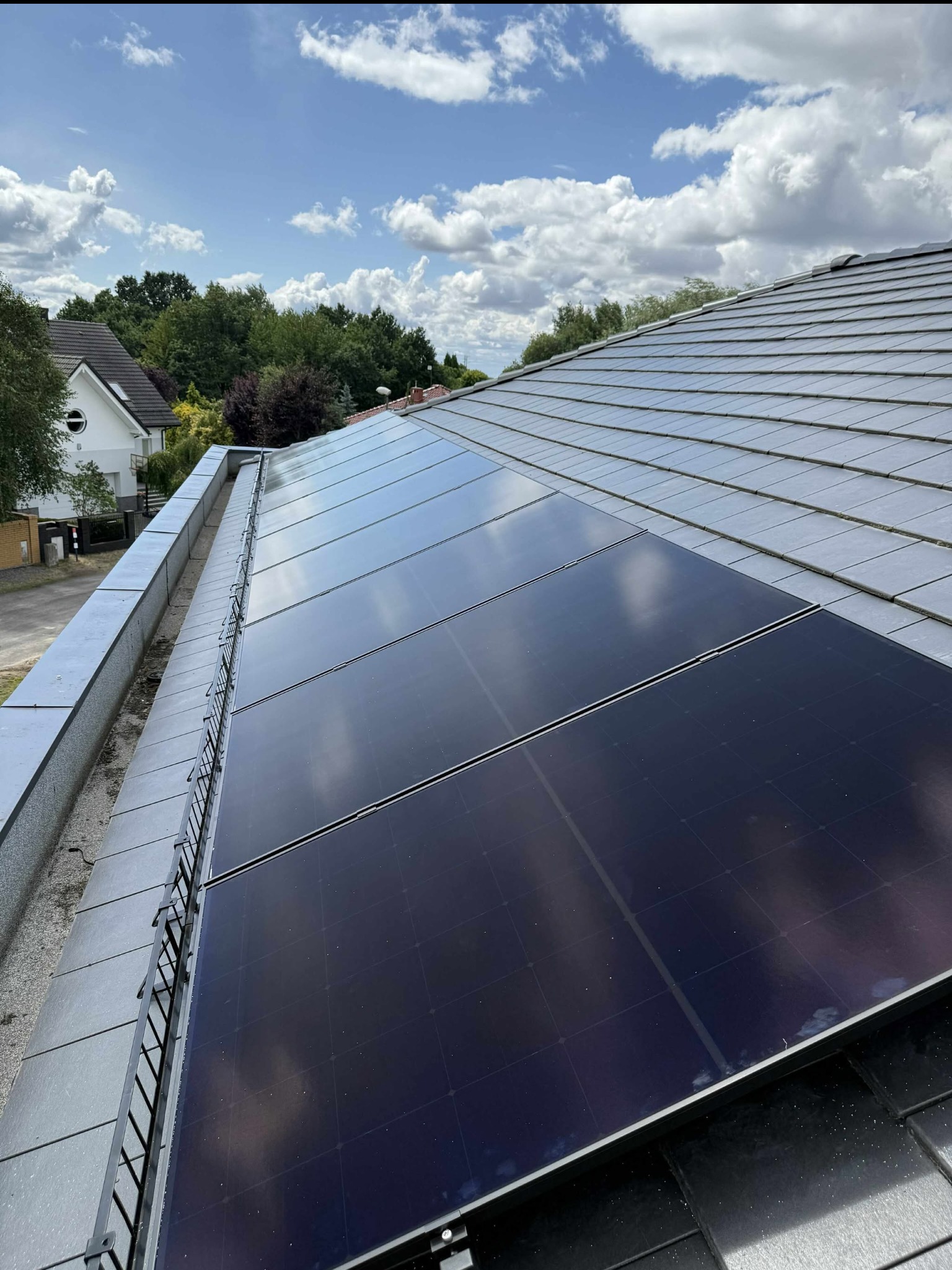 Panele słoneczne na szarym dachu domu w słoneczny dzień. Widok z góry, z widocznymi mocowaniami i elementami konstrukcyjnymi. Tło: błękitne niebo z chmurami i zieleń drzew.