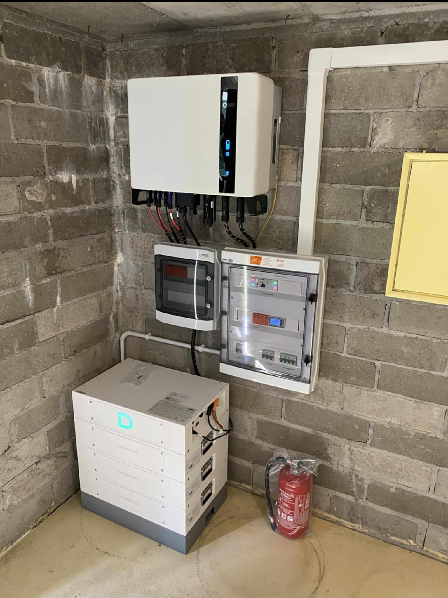 Kompletna instalacja fotowoltaiczna w piwnicy w Szczecinie: falownik, rozdzielnica i akumulatory. Widoczna gaśnica obok akumulatorów.