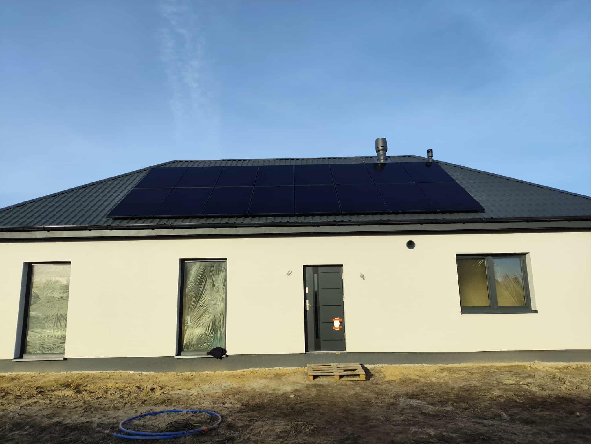 Dom jednorodzinny z panelami słonecznymi na dachu. Okna zabezpieczone folią malarską, teren wokół domu częściowo zagospodarowany. Widoczne palety i rura.