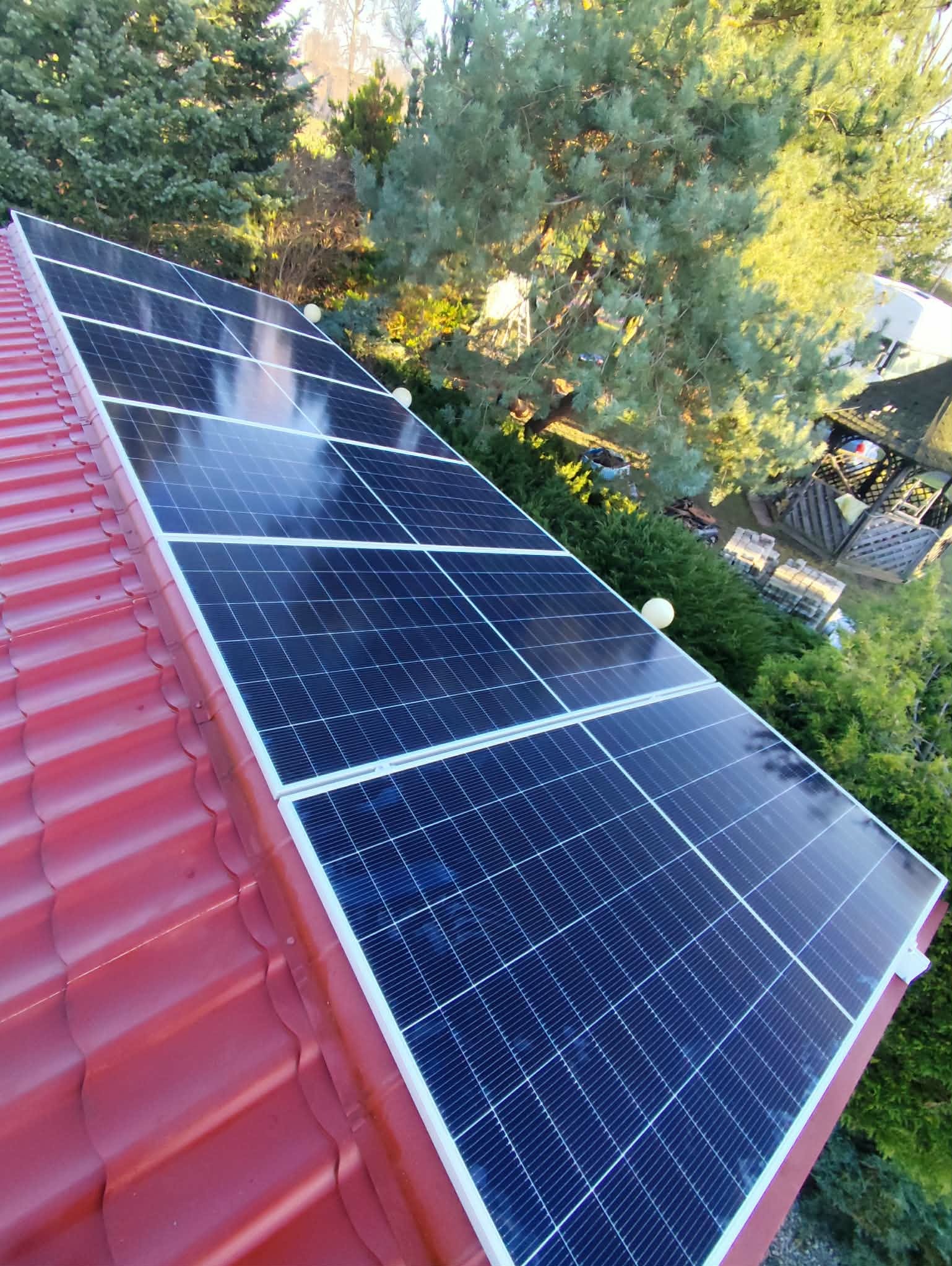 Panele słoneczne na czerwonym dachu, widok z góry, otoczone zielenią drzew i krzewów. Błyszcząca powierzchnia paneli odbija światło słoneczne.