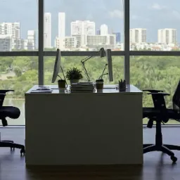 Nowoczesne biuro z dwoma stanowiskami pracy, widok na panoramę miasta przez duże okna, biurka z komputerami, lampka, roślina doniczkowa i krzesła biurowe.