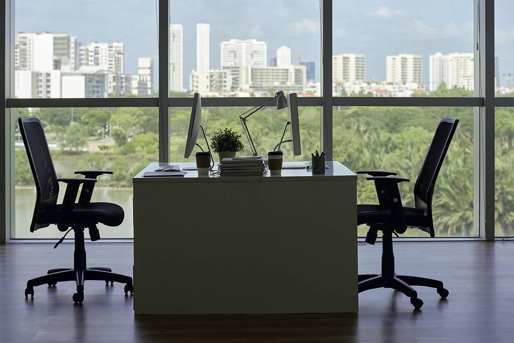 Nowoczesne biuro z dwoma stanowiskami pracy, widok na panoramę miasta przez duże okna, biurka z komputerami, lampka, roślina doniczkowa i krzesła biurowe.