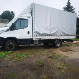 Biały samochód dostawczy z plandeką na błotnistym terenie, widoczne drzewa w tle.