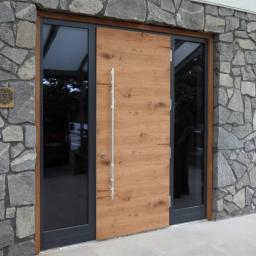 GERLEJ Gerard Lejk - Nowoczesne drzwi wejściowe: drewniane skrzydło z pionowym, metalowym uchwytem, osadzone w czarnej ramie z przeszkleniami, na tle kamiennej elewacji z ozdobną tabliczką.
