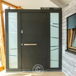GERLEJ Gerard Lejk - Nowoczesne, antracytowe drzwi wejściowe z przeszkleniami, wkomponowane w drewnianą elewację z roletą zewnętrzną, prezentowane w salonie wystawowym.