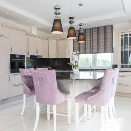 GERLEJ Gerard Lejk - Jasna kuchnia z wyspą i blatem z ciemnego kamienia, oświetlona wiszącymi lampami, z krzesłami w kolorze liliowym na białych nogach.