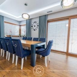 GERLEJ Gerard Lejk - Jadalnia z dużym drewnianym stołem otoczonym niebieskimi, welurowymi krzesłami z białymi nogami, na podłodze panele, duże okna z żaluzjami i zasłonami, na ścianie zegar.