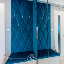 Niebieski, pikowany panel ścienny z wieszakami w przedpokoju, połączony z siedziskiem, obok białe drzwi z przeszkleniami.