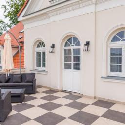 GERLEJ Gerard Lejk - Elegancki taras z szachownicową podłogą, białymi oknami łukowymi i drzwiami, oraz ciemnymi meblami ogrodowymi, z widokiem na czerwoną dachówkę sąsiedniego budynku.