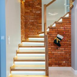 GERLEJ Gerard Lejk - Drewniane schody z podświetleniem LED na stopniach, obłożone cegłą, z dekoracyjnym świątecznym skarpeta na ścianie.