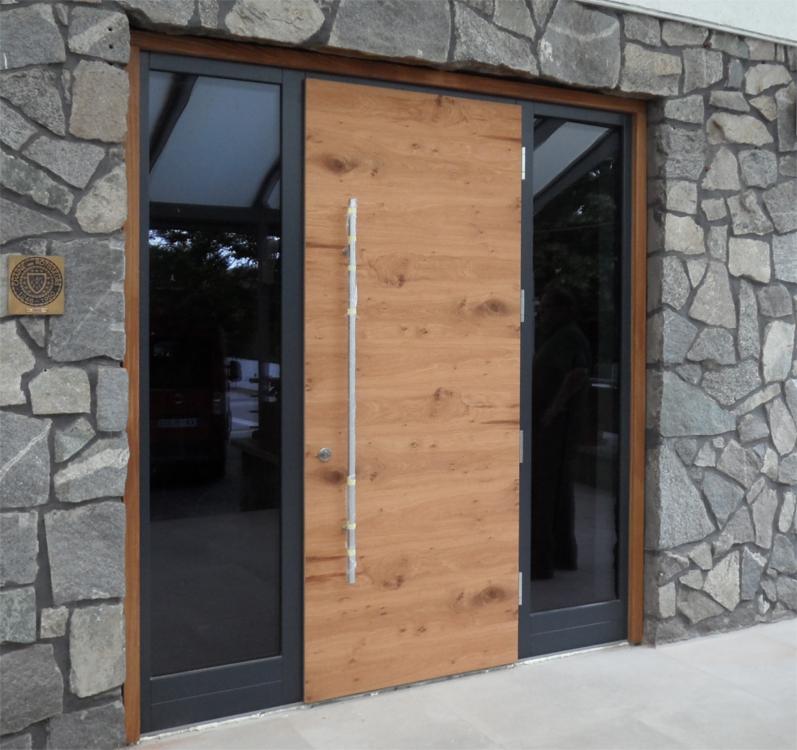 Nowoczesne drzwi wejściowe: drewniane skrzydło z pionowym, metalowym uchwytem, osadzone w czarnej ramie z przeszkleniami, na tle kamiennej elewacji z ozdobną tabliczką.