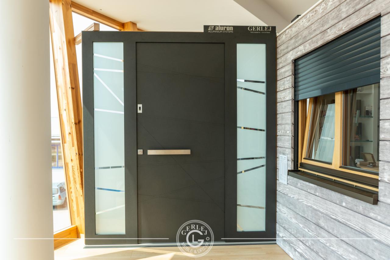 Nowoczesne, antracytowe drzwi wejściowe z przeszkleniami, wkomponowane w drewnianą elewację z roletą zewnętrzną, prezentowane w salonie wystawowym.