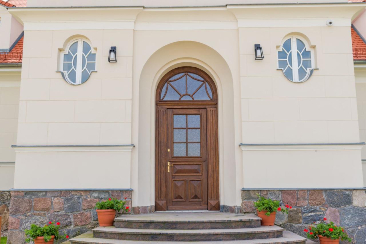 Wejście do budynku z drewnianymi drzwiami z szybami, okrągłymi oknami po bokach i kamienną podmurówką, ozdobione donicami z czerwonymi kwiatami.