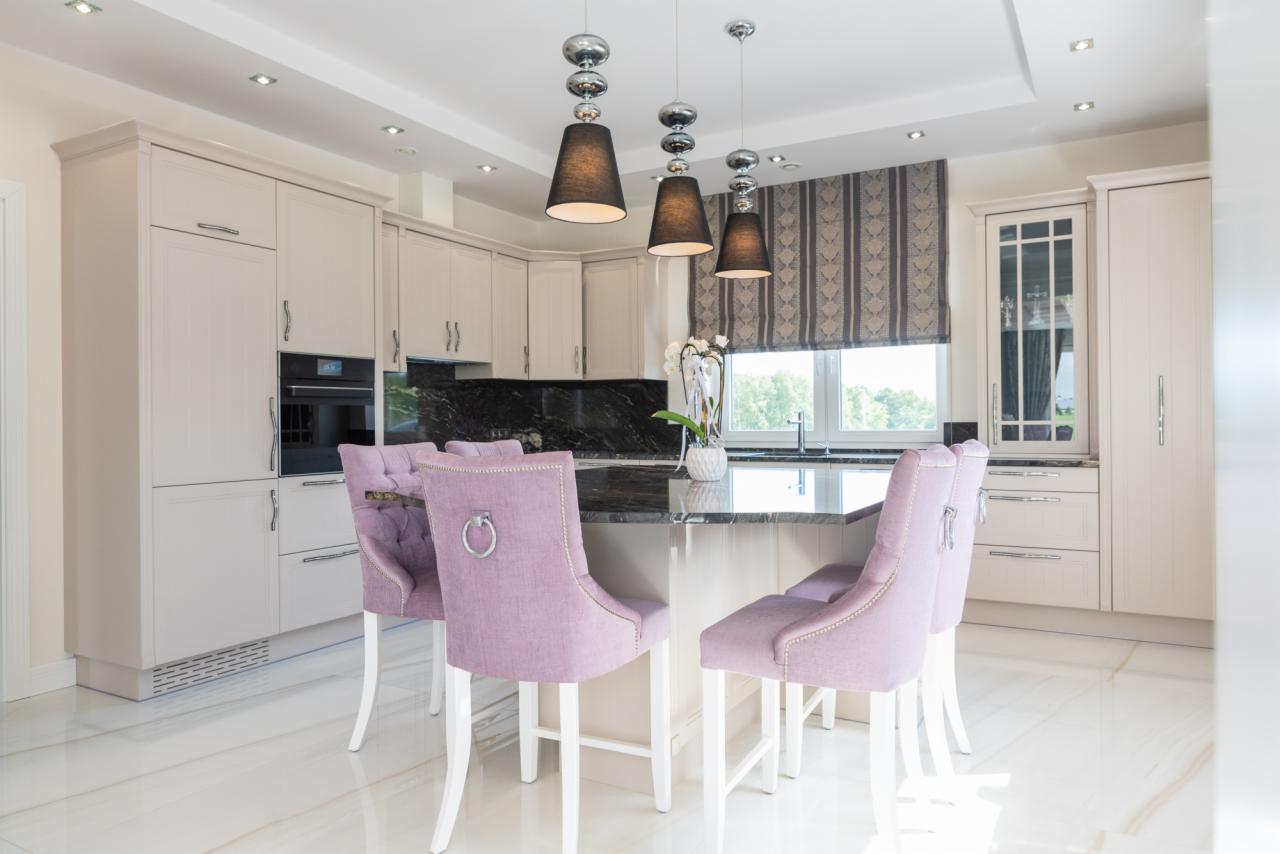 Jasna kuchnia z wyspą i blatem z ciemnego kamienia, oświetlona wiszącymi lampami, z krzesłami w kolorze liliowym na białych nogach.