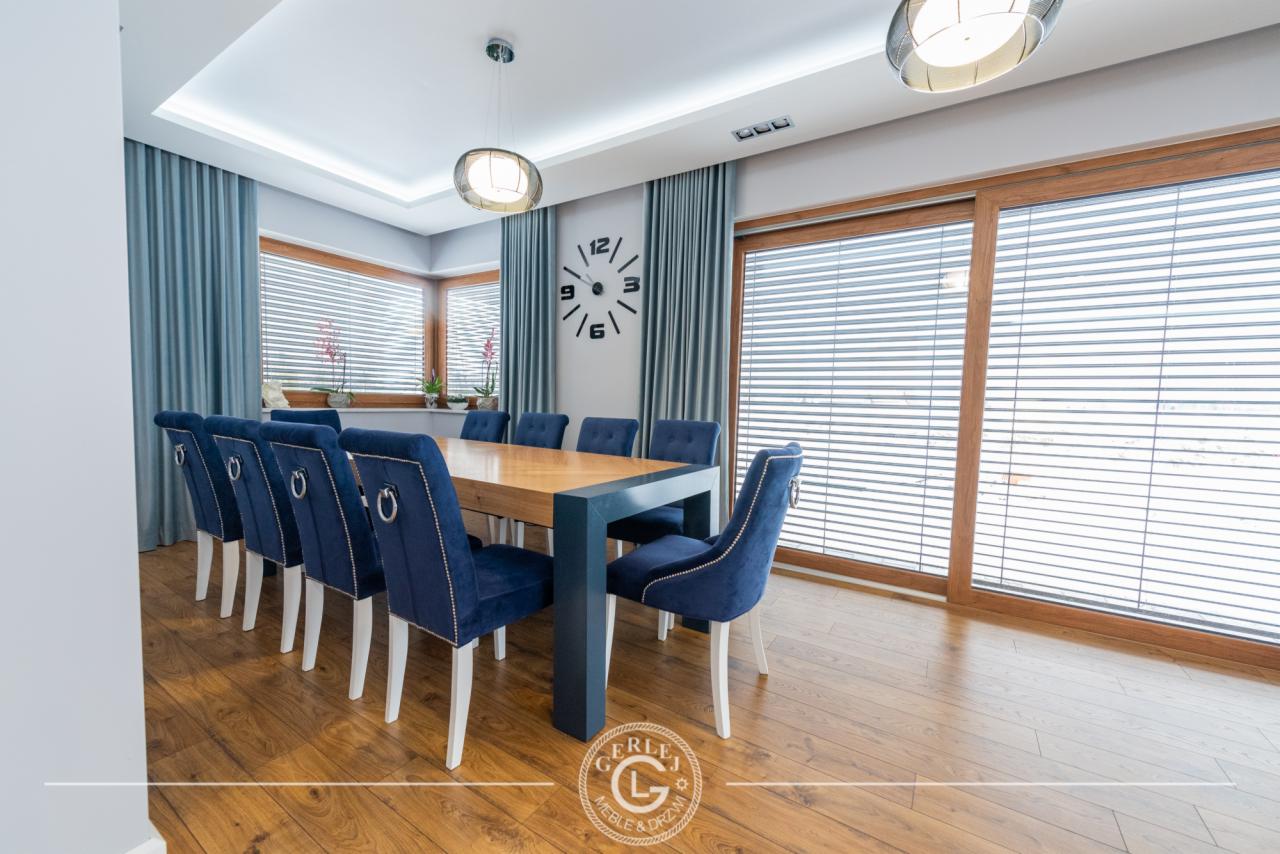 Jadalnia z dużym drewnianym stołem otoczonym niebieskimi, welurowymi krzesłami z białymi nogami, na podłodze panele, duże okna z żaluzjami i zasłonami, na ścianie zegar.