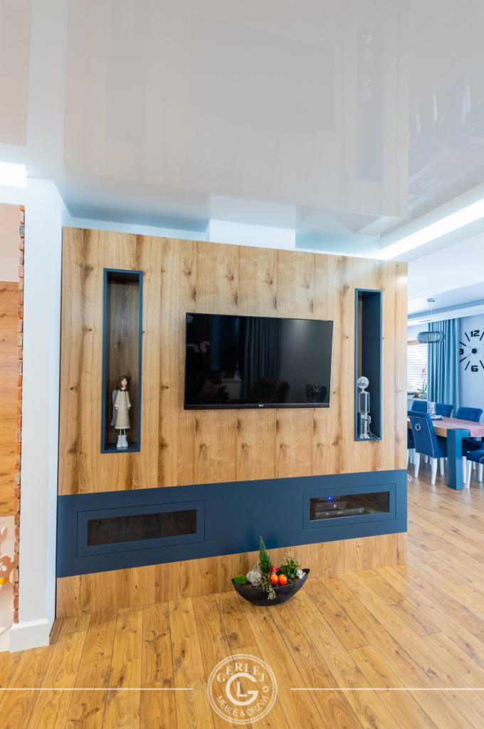 Drewniana ściana telewizyjna na wymiar z wnękami na dekoracje i niebieskim panelem dolnym, z widoczną częścią jadalni w tle.