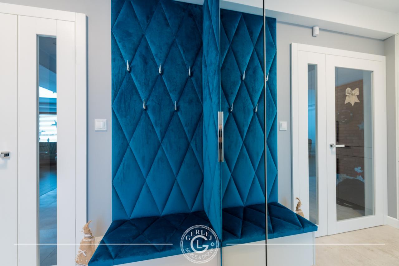 Niebieski, pikowany panel ścienny z wieszakami w przedpokoju, połączony z siedziskiem, obok białe drzwi z przeszkleniami.