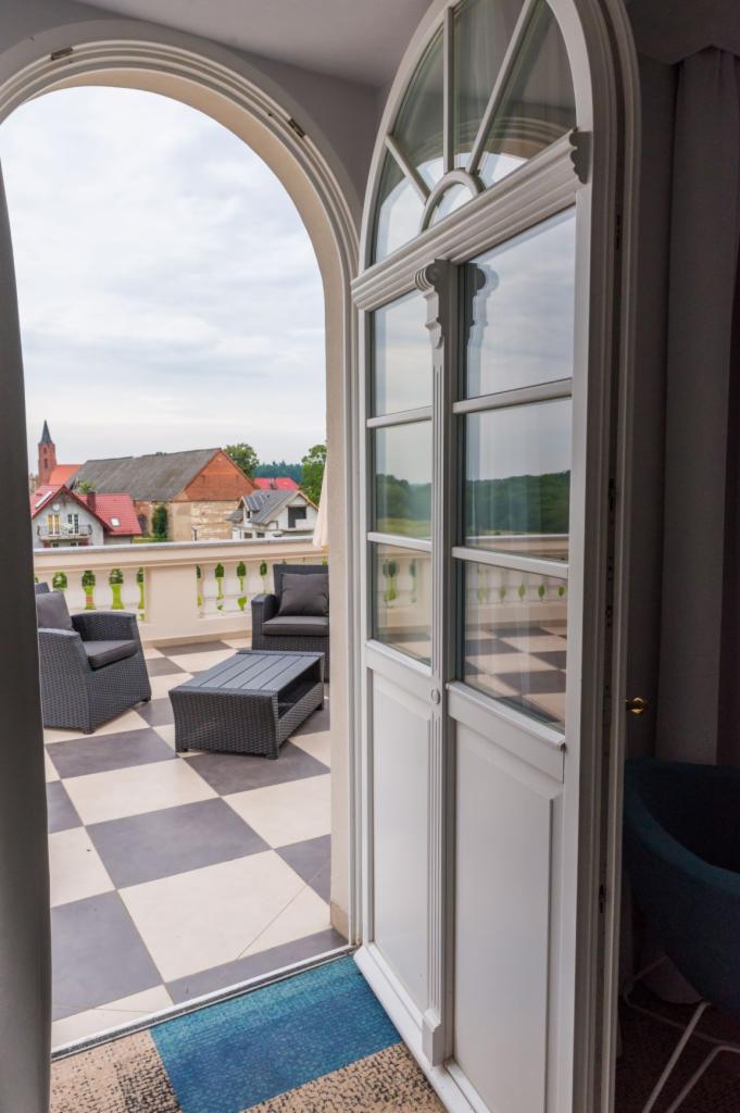 Otwarta, łukowa, biała, szklana konstrukcja drzwiowa z widokiem na balkon z szachownicą na podłodze, szarymi meblami ogrodowymi i widokiem na zabudowania z czerwonymi dachami.