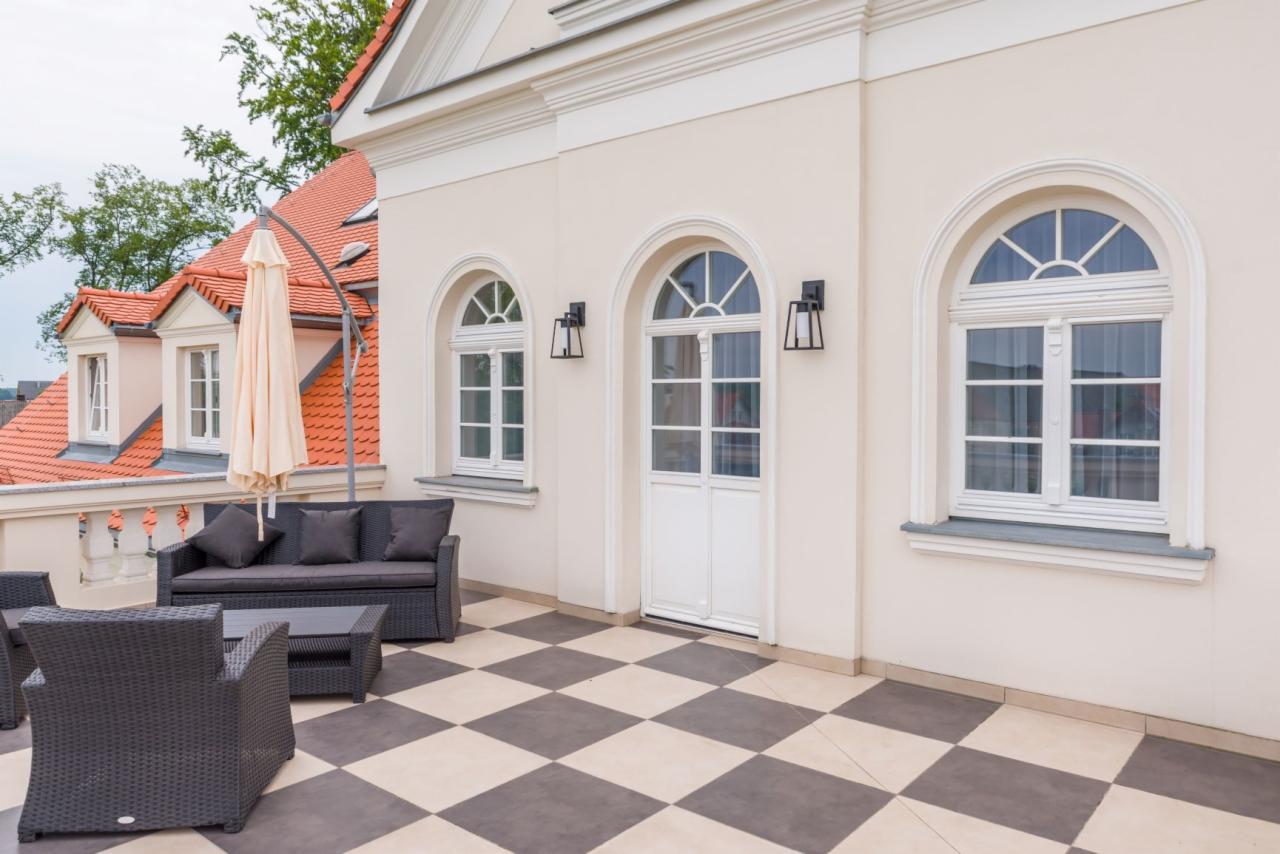 Elegancki taras z szachownicową podłogą, białymi oknami łukowymi i drzwiami, oraz ciemnymi meblami ogrodowymi, z widokiem na czerwoną dachówkę sąsiedniego budynku.