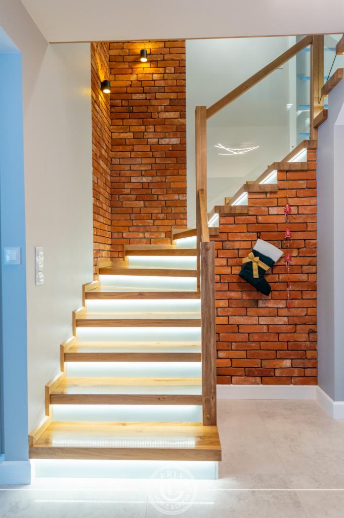 Drewniane schody z podświetleniem LED na stopniach, obłożone cegłą, z dekoracyjnym świątecznym skarpeta na ścianie.
