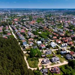 Panoramiczny widok z lotu ptaka na osiedle domów jednorodzinnych w słoneczny dzień, z lasem iglastym po lewej stronie i zabudową miejską w tle.