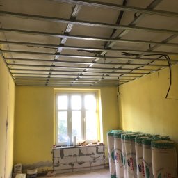 ART-DOM Tomasz Frankiewicz - Metalowa konstrukcja sufitu podwieszanego w trakcie montażu, widoczne przewody elektryczne, żółte ściany, okno z parapetem z bloczków, rolki wełny mineralnej przy ścianie, przygotowania do prac...