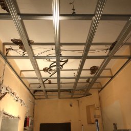 ART-DOM Tomasz Frankiewicz - Montaż metalowej konstrukcji sufitu podwieszanego z widocznymi instalacjami elektrycznymi i uszkodzeniami starego sufitu, ściany w kolorze brzoskwiniowym, drabina w tle.