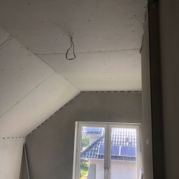 ART-DOM Tomasz Frankiewicz - Poddasze w trakcie wykańczania płytami kartonowo-gipsowymi, widoczne łączenia płyt na skośnym suficie i ścianach, kabel zwisający z sufitu, okno z widokiem na dach z panelami słonecznymi.