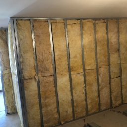 ART-DOM Tomasz Frankiewicz - Montaż ścianki działowej: metalowy stelaż wypełniony wełną mineralną, widoczne profile i izolacja przed płytami gipsowo-kartonowymi.