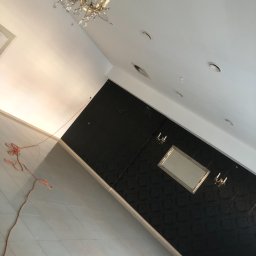 ART-DOM Tomasz Frankiewicz - Wnętrze w trakcie remontu: czarna ściana z wzorem, lustro w złotej ramie, kinkiety, widoczna część sufitu z lampą i kabel elektryczny na podłodze z jasnych płytek.