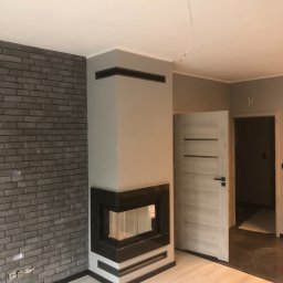 ART-DOM Tomasz Frankiewicz - Wnętrze w Brzegu z kominkiem obudowanym szarą cegłą i czarnymi elementami, widoczne gniazdka elektryczne, biała podłoga i częściowo widoczne drzwi.