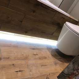 ART-DOM Tomasz Frankiewicz - Ukończona łazienka z drewnopodobnymi płytkami na podłodze i ścianach, podwieszana toaleta, listwa LED pod wanną i fragmentem surowej ściany z cegły.