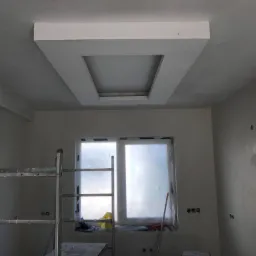 Pokój w trakcie remontu: widoczny sufit z obniżoną, kwadratową konstrukcją z karton gipsu z prostokątnym otworem, ściany w szarym tynku, drabina malarska i okno zabezpieczone folią.